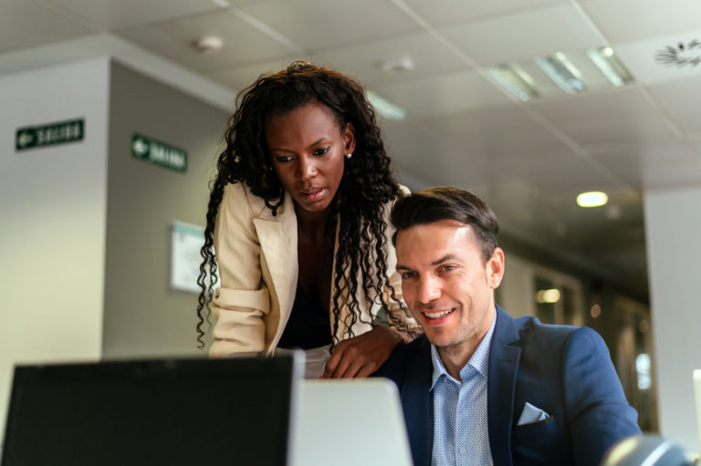 Positive respectable businesspeople including white male and African American female using laptop and reading email with new business offer while working together in modern workplace